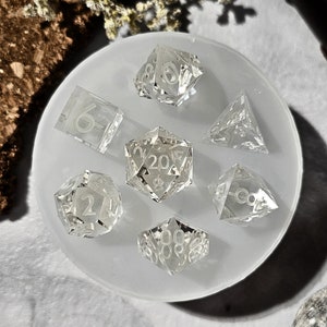 May include: A set of seven clear, glass dice with white numbers. The dice are arranged in a circle on a white silicone mat. The dice include a d4, d6, d8, d10, d%, d12, and d20.