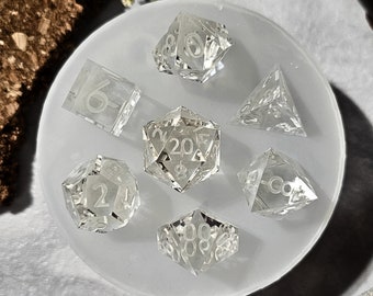 May include: A set of seven clear, glass dice with white numbers. The dice are arranged in a circle on a white silicone mat. The dice include a d4, d6, d8, d10, d%, d12, and d20.