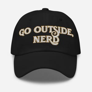 Pode incluir: Boné de beisebol preto com o texto "GO OUTSIDE, NERD" em uma fonte decorativa dourada e branca. O boné tem uma aba curva e uma alça ajustável. Um acessório casual para atividades ao ar livre.