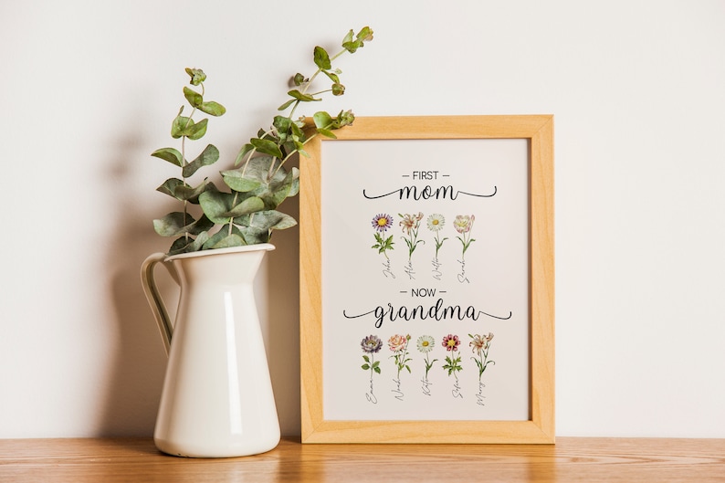 May include: A framed print with a floral design and the text "First Mom - Now Grandma". The print features a row of flowers with names written below each flower. The frame is light brown wood.