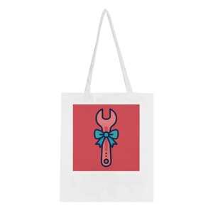 Könnte beinhalten: Weiße Tragetasche mit einem roten Quadrat mit einem Cartoon-Schraubenschlüssel-Design. Der Schraubenschlüssel ist rosa mit blauen und gelben Akzenten und einer blauen Schleife. Die Tasche hat weiße Träger.