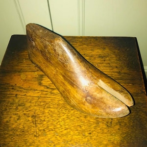 May include: A vintage wooden shoe last, showcasing a foot-shaped form with a split toe. The wood has a warm, brown tone with visible grain, suggesting age and use. The last sits on a wooden surface.