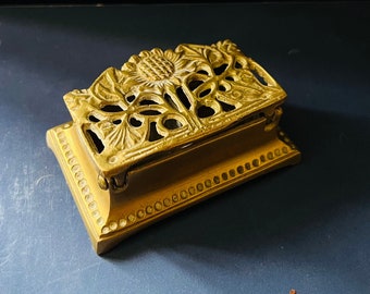 Pode incluir: Uma pequena caixa de latão adornada com um design floral. A caixa tem uma tampa com dobradiças e uma base decorativa.