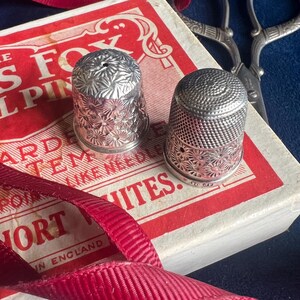 Könnte beinhalten: Zwei silberne Fingerhüte mit filigranen Blumenmustern liegen auf einer rot-weißen Schachtel mit dem aufgedruckten Text "The Fox El Pin" und "Short Whites". Ein rotes Band und eine silberne Schere sind ebenfalls auf dem Bild zu sehen.