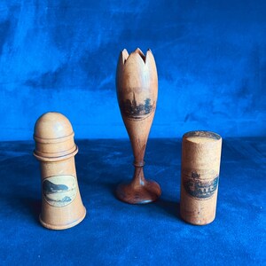 Puede incluir: Tres objetos de madera vintage: un objeto en forma de faro, un jarrón en forma de tulipán y un recipiente cilíndrico. Cada pieza presenta una imagen de paisaje en blanco y negro. Los objetos son de madera clara y están sobre una superficie azul.