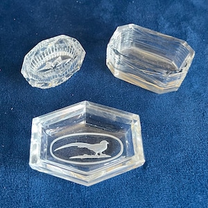 Könnte beinhalten: Drei durchsichtige Glasschmuckschalen. Eine ist oval mit einem Sterndesign, eine andere rechteckig und die dritte sechseckig mit einem Fasan, der in einem Oval eingeätzt ist. Alle liegen auf einer blauen Oberfläche.