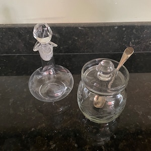 May include: A set of two clear glass containers with etched designs. The larger container has a stopper with a diamond-shaped top. The smaller container has a lid and a small silver spoon inside.