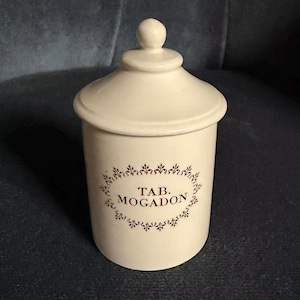May include: A cream-coloured ceramic apothecary jar with a matching lid featuring a knob handle. The front of the cylindrical container displays the text TAB. MOGADON printed in dark brown, enclosed within a decorative, ornate oval border.