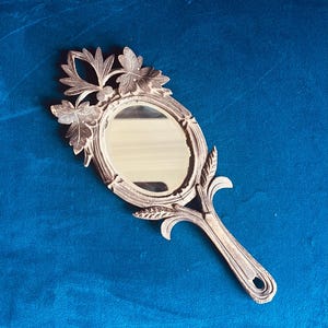 Puede incluir: Un espejo de mano antiguo con una superficie ovalada y un mango decorativo. El marco y el mango son de color plateado y presentan un dise&ntilde;o de hojas y flores. El espejo est&aacute; sobre un fondo azul.