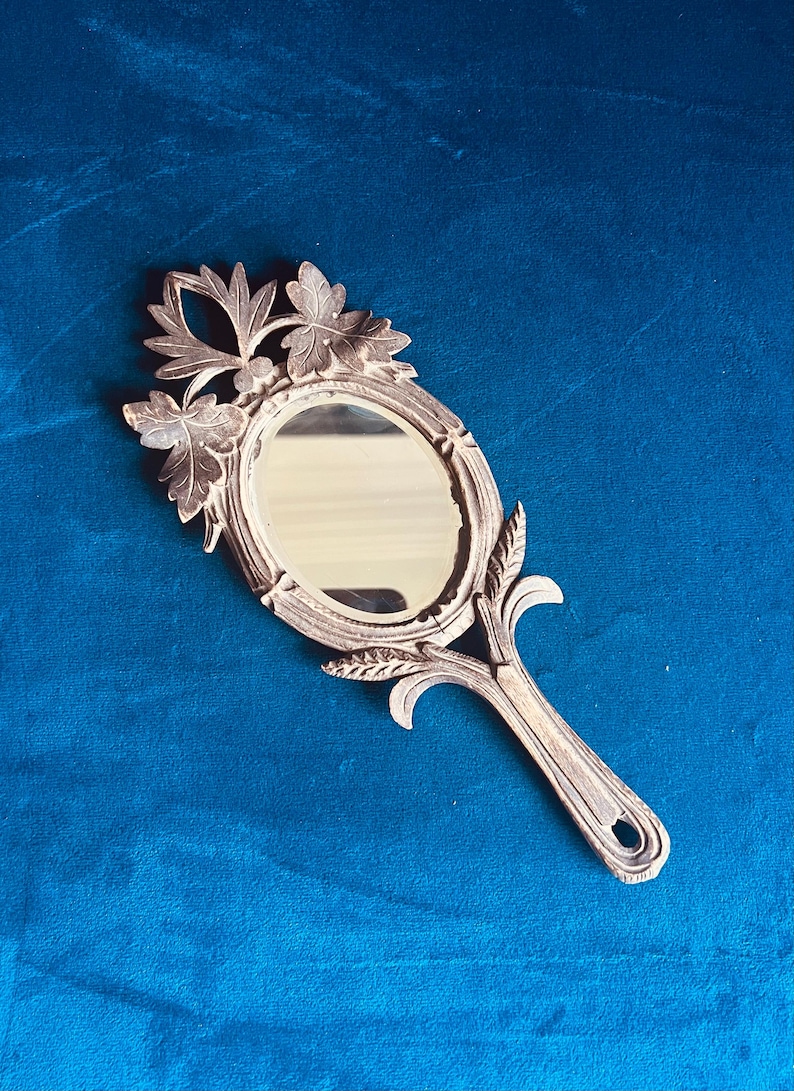 Puede incluir: Un espejo de mano antiguo con una superficie ovalada y un mango decorativo. El marco y el mango son de color plateado y presentan un dise&ntilde;o de hojas y flores. El espejo est&aacute; sobre un fondo azul.