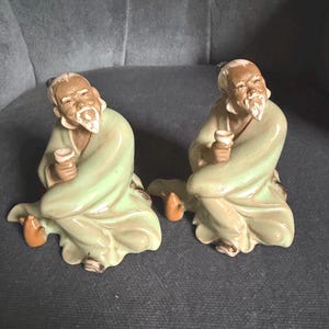 May include: Two ceramic figurines depicting elderly figures with white beards and hair, wearing light green robes. Each figure is seated in a relaxed posture, holding a small white cup in one hand. The glaze has a glossy finish with subtle brown shading.