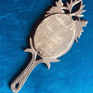 Puede incluir: Un espejo de mano de madera vintage con un espejo ovalado y detalles tallados ornamentados. El mango y el marco presentan intrincados dise&ntilde;os florales y de hojas. El espejo es de color marr&oacute;n claro y descansa sobre una superficie azul.