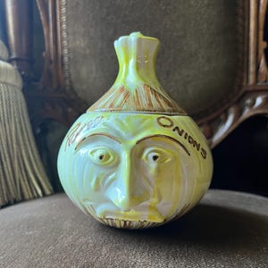 May include: A vintage ceramic onion-shaped container with a lid. The container is painted in shades of yellow and green, with a face design and the word "Onions" written on the side. The lid has brown accents.