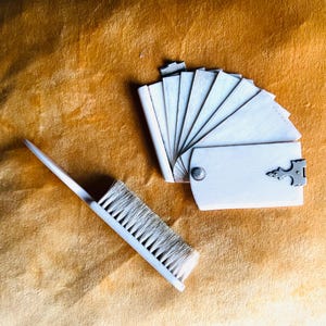 May include: A white brush and a small, hinged fan-shaped object with multiple white panels. The brush has a long handle and bristles. The fan-shaped object has a decorative metal clasp and a metal hinge. The items are on a gold-coloured surface.