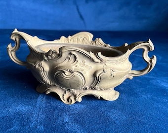 May include: A beige, ornate, antique-style bowl with two handles. The bowl features intricate floral and scrollwork designs. The bowl is made of a light-colored material and sits on a small base. The overall shape is oval.