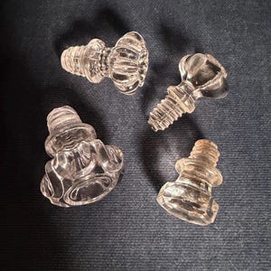 May include: Four clear glass finials with threaded metal-like bases. Each piece features a distinct shape, including fluted, rounded, and faceted designs. These decorative hardware components are transparent and exhibit reflective surfaces throughout.