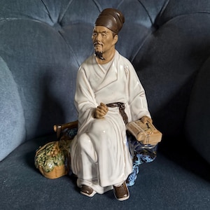 May include: A porcelain figurine of a seated figure wearing a white robe, a dark brown cap, and dark shoes. The figure has a goatee, holds a small rectangular object, and sits beside a woven basket filled with green and brown fruit or foliage.
