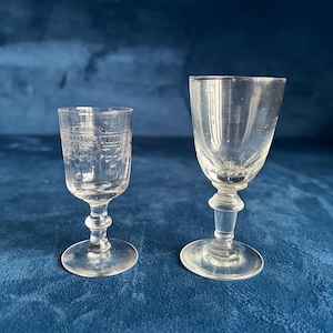 May include: Two clear, stemmed glasses. The glass on the left has a detailed floral pattern etched into the glass. The glass on the right is plain. Both glasses have a round base and a bulbous stem. The glasses are on a blue surface.