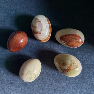 May include: Five smooth, polished, egg-shaped stones. One is solid reddish-brown, one is cream with brown veining, one is translucent with a brown centre, one is solid cream, and one is translucent with green and brown swirls and inclusions.