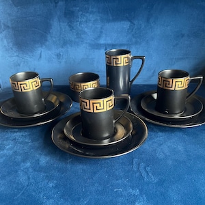 May include: Set of four black and gold teacups and saucers with a geometric pattern. The cups and saucers are arranged on a blue surface.