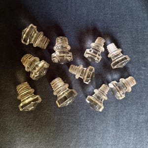 May include: A collection of nine clear, faceted glass knobs, each featuring a hexagonal top and a threaded screw base. These small, transparent hardware pieces are arranged in a scattered pattern, showcasing their geometric shape and reflective surfaces.