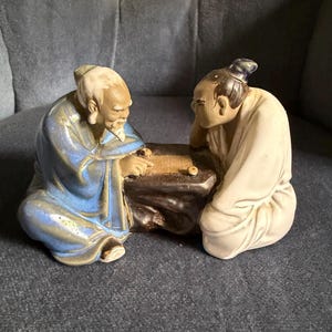 May include: A ceramic figurine depicting two figures sitting on the floor facing each other while playing a board game. One figure wears a blue robe and has a white beard, while the other wears a white robe. They are positioned around a dark brown table.