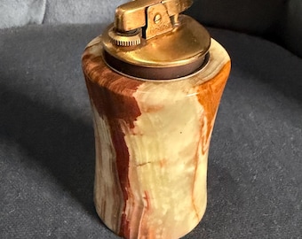 May include: A cylindrical table lighter featuring a polished brass ignition mechanism set into a smooth, variegated stone body with swirling patterns of cream, tan, and deep reddish-brown hues throughout the natural mineral surface.