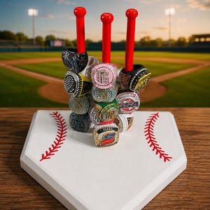 May include: A white home plate with red stitching detail serves as a base for three vertical red baseball bat-shaped posts. Multiple championship rings with various colored stones, metal finishes, and text including USSSA are stacked on the posts.