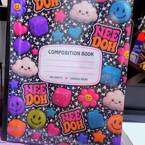 May include: A black composition book with a holographic star pattern and colorful Nee Doh graphics including smiley faces, clouds, hearts, stars, and flowers. The center label reads Composition Book, 100 Sheets, College Ruled, with a small heart icon.