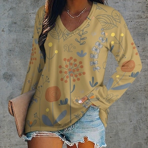 May include: Mustard yellow long-sleeve V-neck shirt with a floral pattern in orange, blue, and yellow. The shirt features a botanical design with various flower and leaf illustrations. The model is wearing denim shorts and holding a clutch.