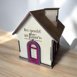 Könnte beinhalten: Eine hölzerne Sparbüchse in Form eines Hauses mit einer pinkfarbenen Tür und einem braunen Dach. Das Haus trägt den Text "Ihr spendet wir flittern" auf der Vorderseite.