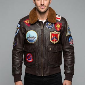 May include: A brown leather bomber jacket with a brown faux fur collar. The jacket features multiple embroidered patches, a zipper closure, and two front pockets. The jacket has a ribbed hem and cuffs.