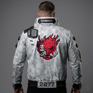 Può includere: Giacca bomber grigio chiaro con dettagli neri e la scritta "SAMURAI" sul colletto. La schiena presenta un design grafico rosso e bianco e il numero "2077".