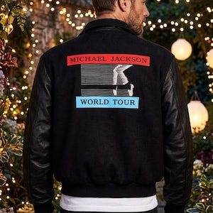 May include: Black bomber jacket with black leather sleeves. The back of the jacket features the text "MICHAEL JACKSON" in red, a graphic of a dancer, and "WORLD TOUR" in blue. The jacket is worn over a white t-shirt.