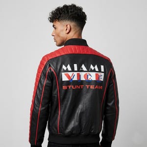 Puede incluir: Chaqueta de cuero negra y roja con el texto "MIAMI VICE STUNT TEAM" en la espalda. La chaqueta tiene un panel acolchado rojo en los hombros y la parte superior de la espalda, con ribete rojo. Cuello y puños negros.
