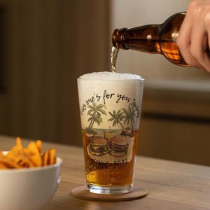 May include: A clear pint glass with a beach scene and the text "This one's for you Jimmy". The glass is filled with beer and foam, with a burger graphic. A bottle pours beer into the glass. A bowl of chips is in the background.