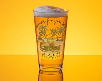May include: A clear pint glass filled with beer and topped with white foam. The glass features a tropical scene with palm trees, burgers, and the text "This one's for you Jim" and "1946-2023". The background is a gradient of yellow.