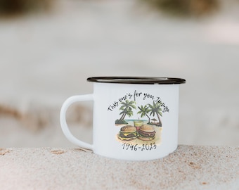 May include: White enamel mug with a black rim and handle. The mug features a watercolor illustration of two burgers on a beach with palm trees and the text "This one's for you Jimmy" and the years 1946-2023.