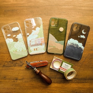 May include: Four clear phone cases with different designs. The cases feature a sunset, a moon, a train, and a landscape. The cases are on a wooden surface.
