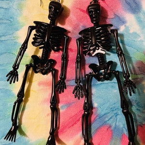 May include: A pair of black skeleton earrings. The skeletons are posed with their arms outstretched and their legs crossed. The earrings are hanging from a tie-dye background.