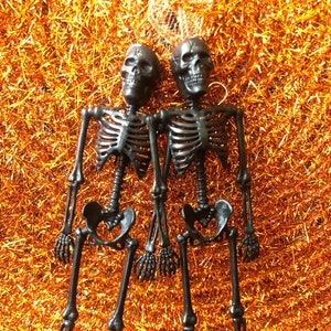 May include: Two black plastic skeleton decorations, perfect for Halloween. Each skeleton has a detailed skull, rib cage, and articulated limbs. They are posed together against a backdrop of orange tinsel.
