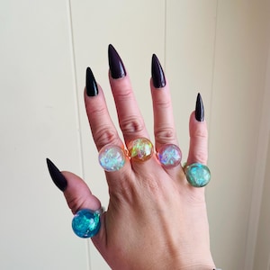 May include: Five rings featuring spherical, translucent resin settings, each containing iridescent, shimmering flakes in distinct colors including blue, pink, orange, and teal, worn on the fingers of a hand with dark, pointed fingernails.