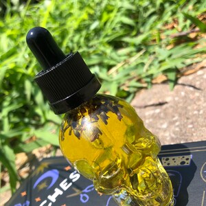 Peut inclure: Une bouteille en verre transparente en forme de cr&acirc;ne remplie d'un liquide jaune et d'herbes s&eacute;ch&eacute;es. La bouteille a un bouchon compte-gouttes noir.