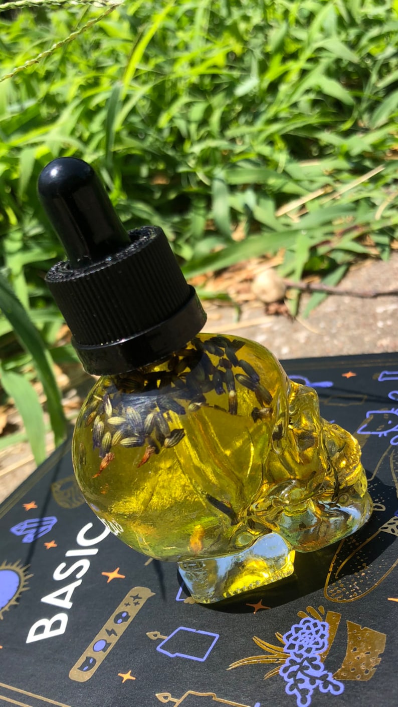 Peut inclure: Une bouteille en verre transparente en forme de cr&acirc;ne remplie d'un liquide jaune et de fleurs de lavande s&eacute;ch&eacute;es. La bouteille est munie d'un bouchon compte-gouttes noir.
