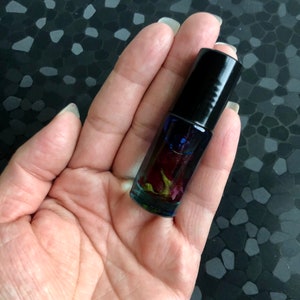 Peut inclure: Un petit flacon en verre avec un bouchon noir rempli d'un liquide clair et de p&eacute;tales de rose s&eacute;ch&eacute;es. Le flacon est tenu dans une main.