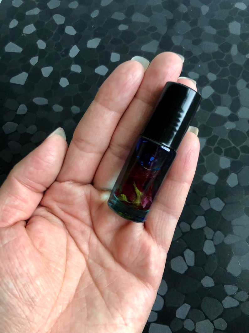 Peut inclure: Un petit flacon en verre avec un bouchon noir rempli d'un liquide clair et de p&eacute;tales de rose s&eacute;ch&eacute;es. Le flacon est tenu dans une main.