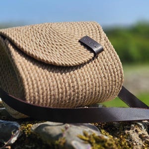 Peut inclure: Un sac à bandoulière beige tissé avec une fermeture à rabat et une bandoulière en cuir marron foncé. Le sac a un aspect texturé en forme de corde et une petite fermeture à languette en cuir marron foncé. Le sac est posé sur une surface en pierre.