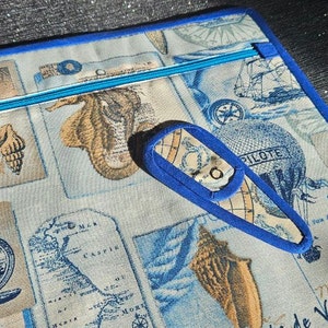 May include: A square, light blue and beige travel-themed zippered pouch with a matching bookmark. The pouch features a blue zipper and trim, and a pattern of vintage maps, compasses, and hot air balloons. The bookmark has a loop.