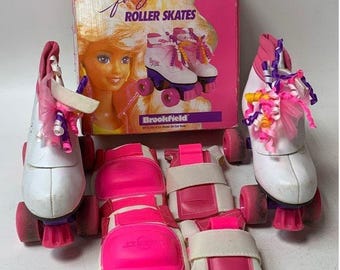 May include: A set of white roller skates with pink wheels and purple accents. The skates have pink and purple ribbons and bows. The set also includes pink and white knee pads and elbow pads. The box says "Barbie for girls roller skates" and "Brookfield".