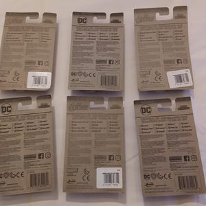 May include: Six toy action figure packages with brown cardboard backing. The packages feature the logos for DC and Marvel comics, with text and barcodes visible. The packages are arranged on a white surface.